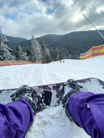 Woman in ski suit lying on snow with snowboard and beautiful view from the top of the Carpathian Mountains to the snowy mountains and fir trees on background, first person photo, ski trail in winter Bukovel, Ukraineの写真素材