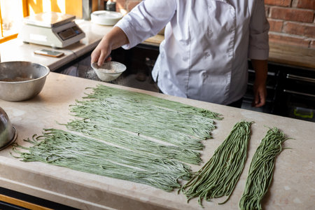 woman sprinkles dough with sifted flour. Cook preparing handmade pasta. Womans hands working with dough to make pasta. Chef making green dough blanks to make craft pasta. A variety of spinach dough preparations lie on a white kitchen table. Chef in white jacket preparing food in restaurant kitchen.の写真素材
