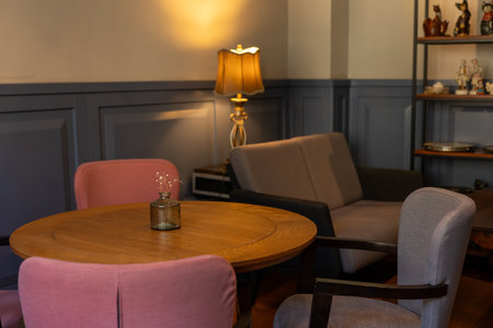 A cozy restaurant room is highlighted by a round wooden table surrounded by pastel-colored chairs. A soft lamp casts a warm glow, enhancing the relaxed atmosphere. A gray couch sits nearby, while a shelf filled with decorative objects showcases personal touches. The serene environment suggests a perfect spot to unwind in the afternoon.の写真素材