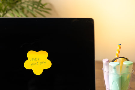 A bright yellow sticky note with handwritten text saying Have a nice day is placed on a laptop screen, creating a cheerful atmosphere. Next to the laptop, a clear container holds colorful markers and pens. The wooden desk is complemented by a green plant in the background, enhancing the inviting workspace vibe.の写真素材