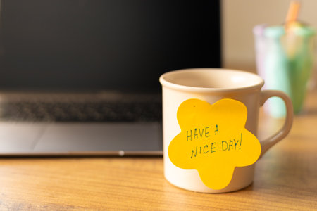A mug sits on a wooden desk next to a laptop, featuring a bright yellow sticky note with the message Have a nice day. The warm atmosphere of the workspace suggests a positive start to the day. Natural light softly illuminates the scene, creating an inviting and motivational setting for work or study. High quality photoの写真素材