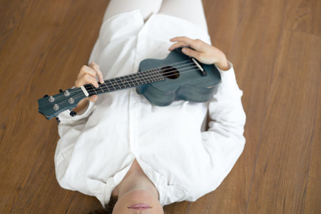 The individual enjoys a moment of tranquility while lying on a polished wooden floor, strumming a ukulele. Dressed in a white shirt, they exhibit a sense of calm and creativity. The indoor setting offers a warm ambiance, encouraging relaxation and musical expression during their leisurely time.の写真素材