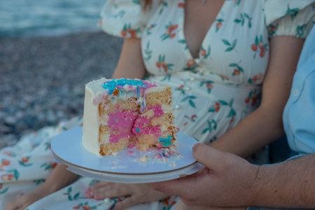 A joyful family gathers by the riverside for a gender party. A small cake decorated with pink and blue colors, sits on a blanket surrounded by fresh fruits. The parents prepare to cut the cake, while their toddler eagerly reaches out. The scene depicts warmth, love, and togetherness during a sunny afternoon.の写真素材
