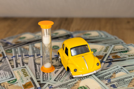 A yellow toy car is positioned next to an hourglass on a stack of hundred-dollar bills. This arrangement conveys a theme of time and money, highlighting their relationship.の写真素材