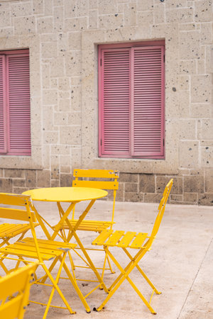 A cozy outdoor setting features vibrant yellow chairs and a table against a textured stone wall with charming pink shutters. The scene captures a relaxed and inviting atmosphere. Positive atmosphereの写真素材