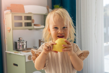 Cute adorable blonde toddler girl eating fresh pear . Hungry happy baby child of two year holding fruit. Toy kitchen in living room on background. Lifestyle and parentship conceptの写真素材