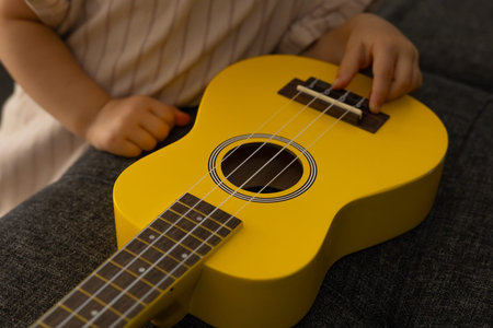 A toddler in a light outfit joyfully strums a bright yellow toy guitar or ukulele while standing on...