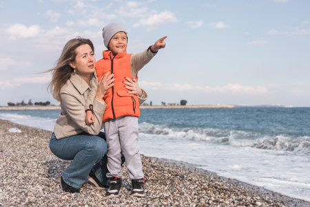 A mother and her young son stand together on a pebbled beach during winter. They smile at each other while the waves lap gently at their feet, and the sky is clear with soft clouds. Family lifestyleの写真素材
