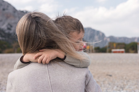 Close-up mother holds her young son close while standing on a quiet beach during winter. The gentle breeze blows through her hair as they enjoy their time together against a mountainous landscape.の写真素材