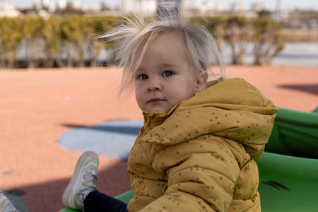 A young child toddler girl with tousled blonde hair sits on a green slide in a playground. The sunny day highlights the vibrant play area, creating a cheerful atmosphere. Winter in Antalya, Turkeyの写真素材