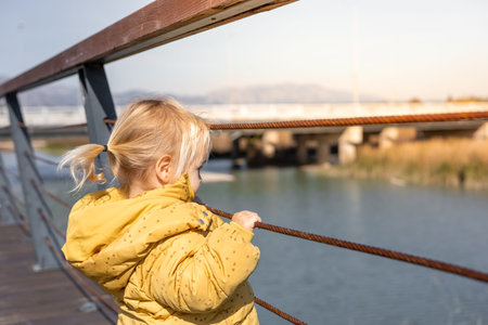 A young child stands on a wooden platform, gazing out at a calm river and lush grasslands. Dressed...