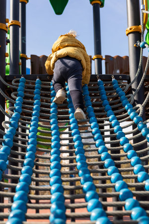 A young child in a yellow jacket climbs a blue and black rope structure at a playground. The sun...