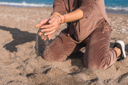 A person in casual brown attire kneels on a sandy beach, allowing grains of sand to slip through their fingers. The bright blue ocean in the background enhances the serene atmosphere.の写真素材