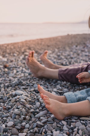 Family enjoys peaceful moment by the beach, with adults and children stretching their bare feet on the smooth pebbles. The gentle waves and soft sunset create a serene atmosphere. Legs without shoesの写真素材