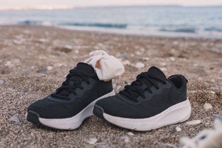 Pair of black female sneakers sits on the warm sand on beach. Sun sets over the calm ocean or sea, casting a golden glow, suggesting relaxation and leisure after sport training, gym, fitness, runningの写真素材