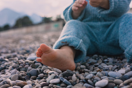 A young girl child toddler sits comfortably on a pebbled beach, showcasing bare feet and a cozy outfit. The tranquil ocean serves as a backdrop under a clear sky, creating a peaceful atmosphere.の写真素材