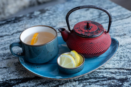 A tea setup features a traditional teapot and a blue cup filled with warm herbal mint tea and lemon slices. Nearby, a small dish holds additional lemon pieces on a granite surface.の写真素材