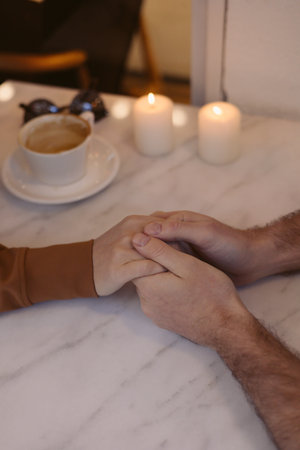 Two lovers hands clasp each other warmly at small table in a cafe. Candles glow softly in the...
