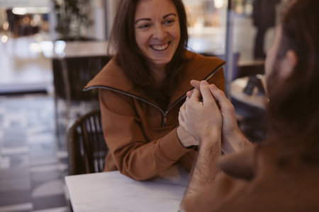 Portrait of woman smiles brightly while touching and holding male hands, sitting with a man at a cafe. The atmosphere is warm and inviting with natural light pouring through the windows.の写真素材