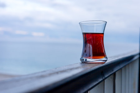 A clear glass filled with turkish black tea rests on a wooden balcony railing. In the background, a calm ocean or sea reflects a muted gray sky, creating a serene relax atmosphere.の写真素材