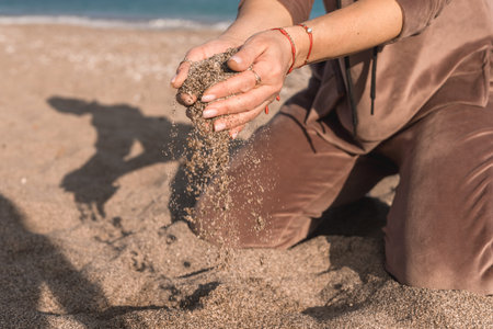 A woman seated on a sandy beach is playfully sifting sand through her fingers. The gentle ocean waves are visible in the background under a clear sky, creating a serene atmosphere.の写真素材