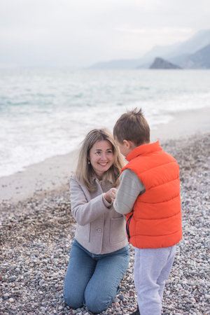 Joyful moment at the beach as a mother kneels to connect with her son on a winter day. They are surrounded by pebbles and the calming waves of the sea in the background. Family fun and rest togetherの写真素材
