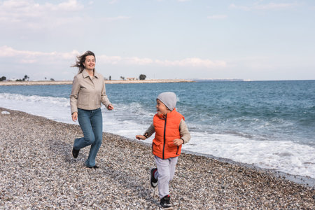 Mother and her son boy run on a pebble beach during winter. Boy wears a bright orange vest and a gray hat, while the mother enjoys the chilly air as they play near the ocean or seaの写真素材