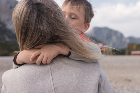 Close-up mother holds her young son close while standing on a quiet beach during winter. The gentle breeze blows through her hair as they enjoy their time together against a mountainous landscape.の写真素材