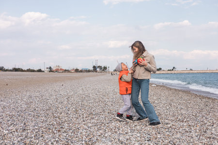 A mother and her son play together on a pebble-strewn beach during winter. They are laughing and...