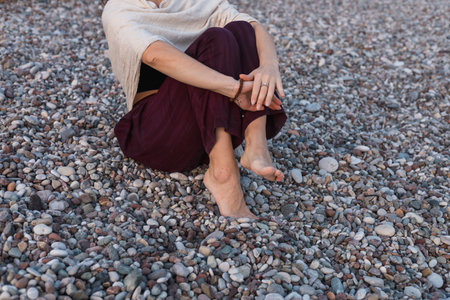 A woman relaxes on a pebble beach, wrapping herself in a light shawl. She sits with her legs feet drawn close, taking in the view of gentle waves and tranquil surroundings as the sun sets.の写真素材