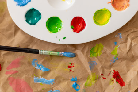 A palette filled with bright paint sits beside a used paintbrush. Colorful strokes and splatters cover the surface. Painting with children, family hobby, leisure activity.の写真素材