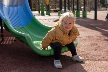 A young toddler child girl wearing a yellow jacket joyfully slides down a slide at a playground. The setting is filled with three vibrant slides under clear blue skies, perfect for outdoor fun.の写真素材