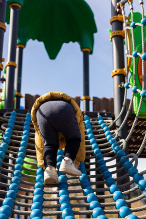 A young child in a yellow jacket climbs a blue and black rope structure at a playground. The sun shines brightly, creating a cheerful atmosphere perfect for outdoor play.の写真素材