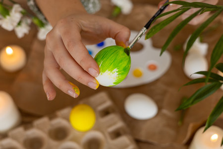 A person decorates an egg with bright green paint among candles and a palette. The setting features...