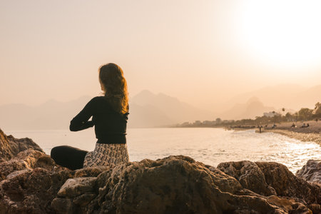 Yogi woman sits peacefully on a rocky shoreline, practicing meditation as the sun sets over the horizon. The soft colors of dusk reflect on the calm water, creating a tranquil atmosphere.の写真素材