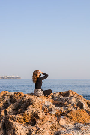 A woman sits on a rock formation by sea or ocean, gazing into the distance. The bright sun shines down on the peaceful water, creating a serene atmosphere. The coastline is visible in the background.の写真素材