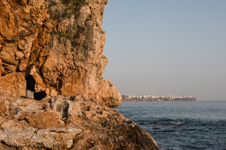 A person sits peacefully on a rocky shore, meditating as dawn breaks. The calm sea reflects the soft light, while a coastal city skyline is visible in the background.の写真素材