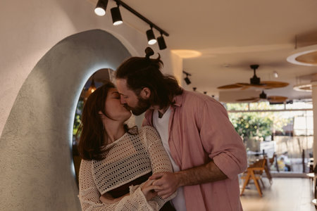 A couple leans in for a tender kiss inside a stylish cafe filled with light. Their affectionate moment captures the warmth of love and connection in a cozy urban space.の写真素材