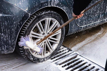 A person uses a brush to clean a car wheel at a detailing center. The wheel is surrounded by soap bubbles and water, with bright daylight illuminating the setting. Self-service car wash processの写真素材