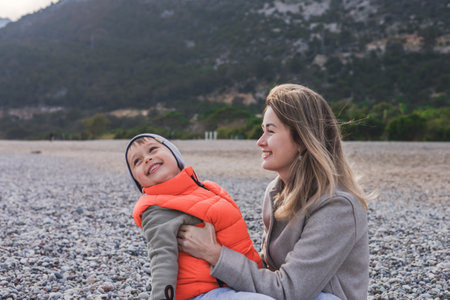 On winter day, a mother and her son enjoy a heartwarming moment at a pebble beach. Kissing and touching by noses. The tranquil sea and distant mountains create a serene backdrop for their affection.の写真素材