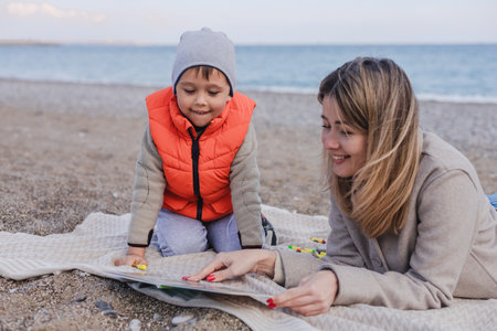 A mother and her son enjoy a winter day on the beach as they read a book together. The tranquil sea and pebbled shore create a serene backdrop for their bonding moment.の写真素材