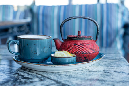 A red teapot sits beside a blue cup on a marble table at a cozy cafe. A small dish holds lemon...