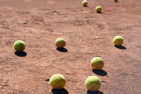 A collection of tennis balls rests on a dusty clay court under bright sunlight. The scene captures...