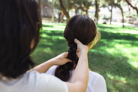 A woman is carefully braiding her daughters hair while sitting outdoors in a lush green park surrounded by trees. The sunlight filters through the leaves, creating a serene atmosphere.の写真素材