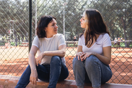 Two women mother and daughter sit on concrete ledge, engaging in cheerful conversation while enjoying their time at a tennis court. Lush greenery surrounds the area, creating a relaxed atmosphere.の写真素材