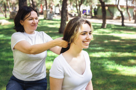 In a lush green park, two women mother and daughter share lighthearted moment. One styles the others...