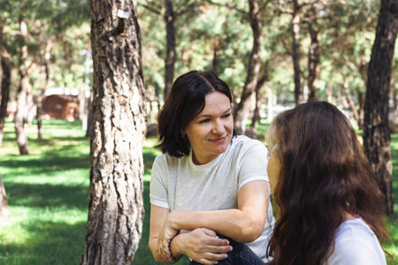 In a lush green forest, two women mother and daughter are seated on the ground, immersed in conversation. Sunlight filters through the trees, creating a calm and relaxed atmosphere.の写真素材