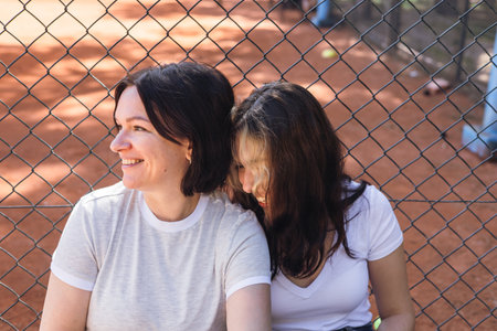 Two woman mother and teenage daughter sit side by side, sharing smiles and laughter in a vibrant park. The warm sun shines down, highlighting their casual outfits and relaxed demeanor.の写真素材