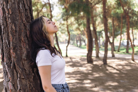 Beautiful young woman leans back against tree, eyes closed, savoring peaceful atmosphere of a sunny park. Sunlight filters through the leaves, creating a serene moment in nature. Merging with natureの写真素材