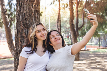 In vibrant park filled with trees, two women, other and teenage daughter pose together for selfie. One has long, wavy hair while the other smiles brightly, both enjoying leisure moment in sunny dayの写真素材